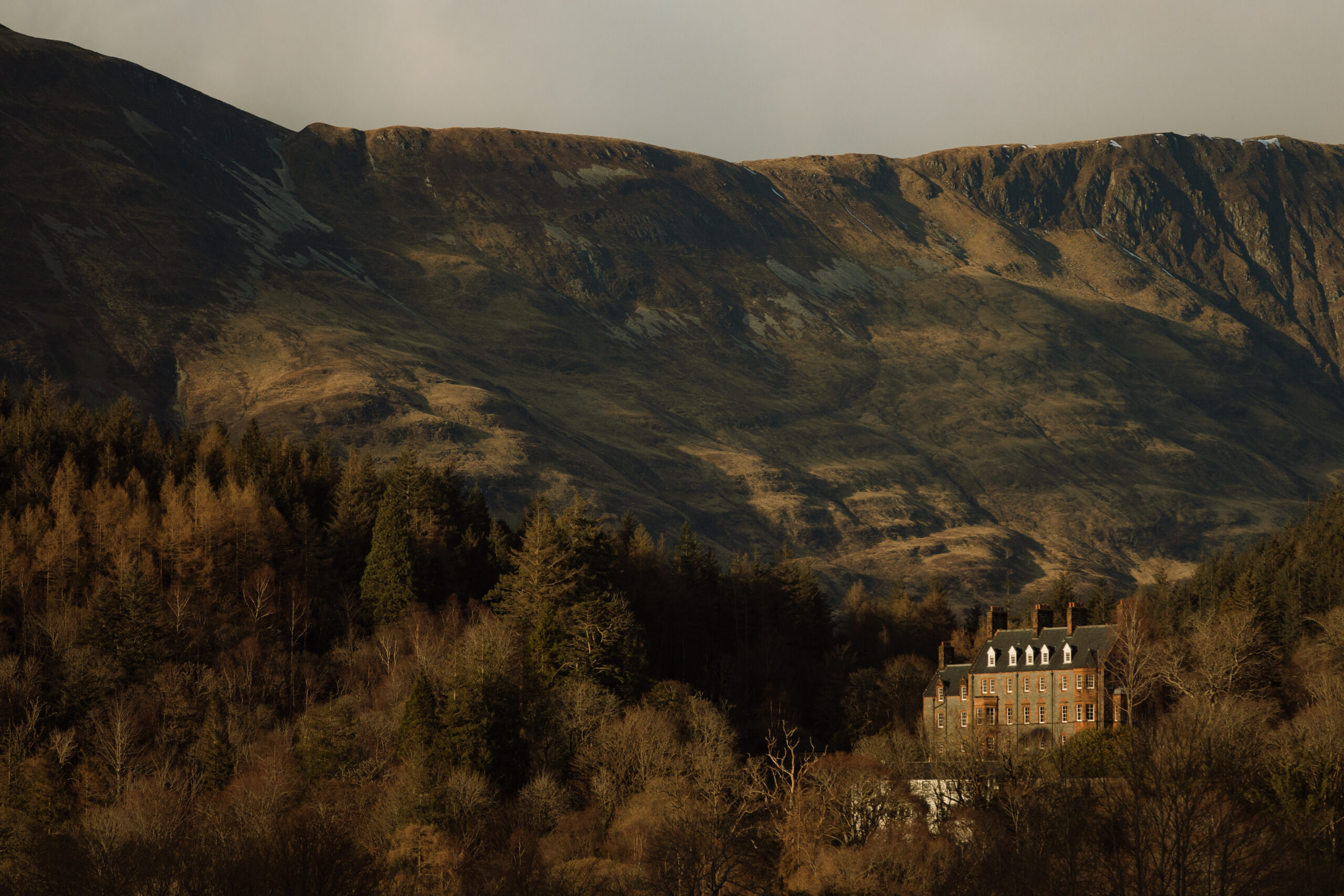 A stone house among autumn trees beneath a Highland ridge in low sun